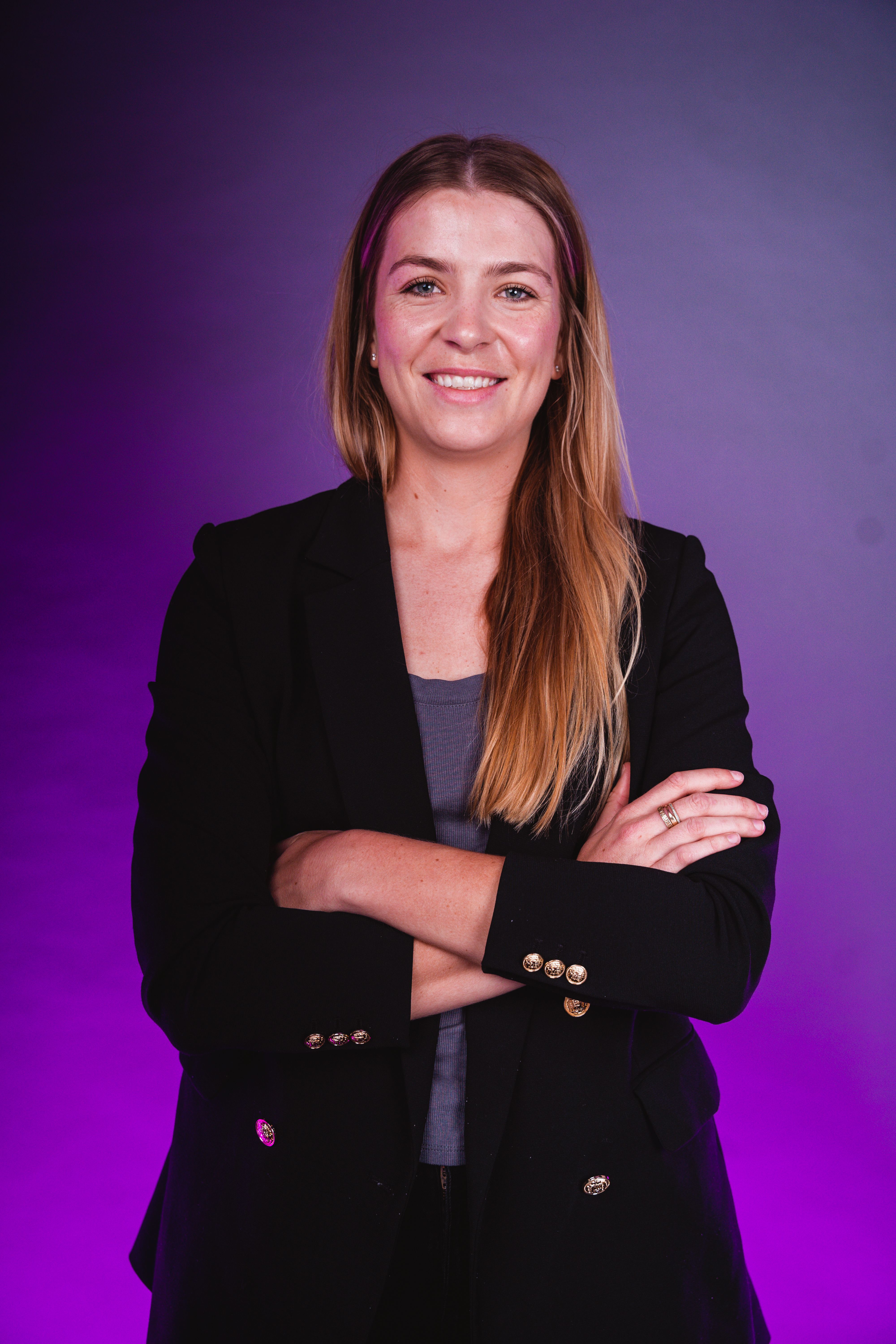 A woman stands with her arms crossed in a black blazer on a purple background