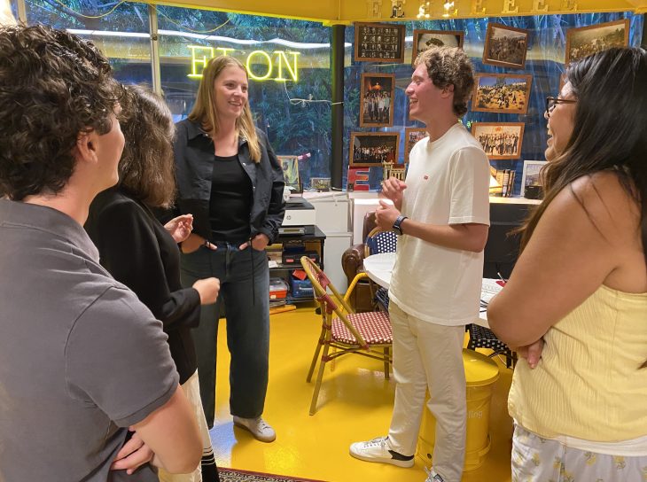 A woman in a jean jacket and jeans talks with a group of people. An illuminated Elon sign is behind them