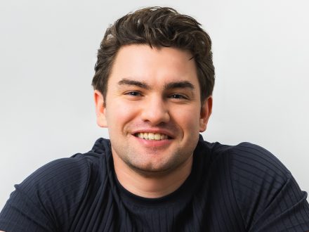 A man in a black shirt poses for a photo on a white background smiling