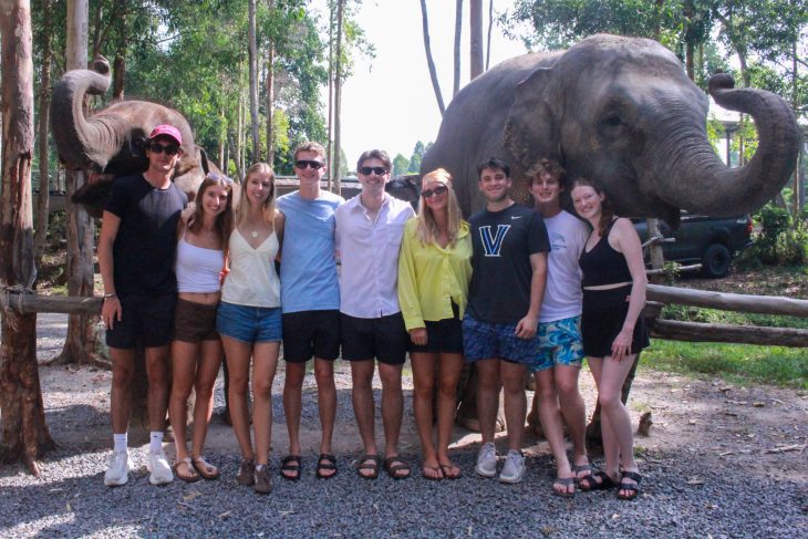 A group of Business Fellows poses outdoors with two elephants standing behind them during the Asia Pacific semester experience.