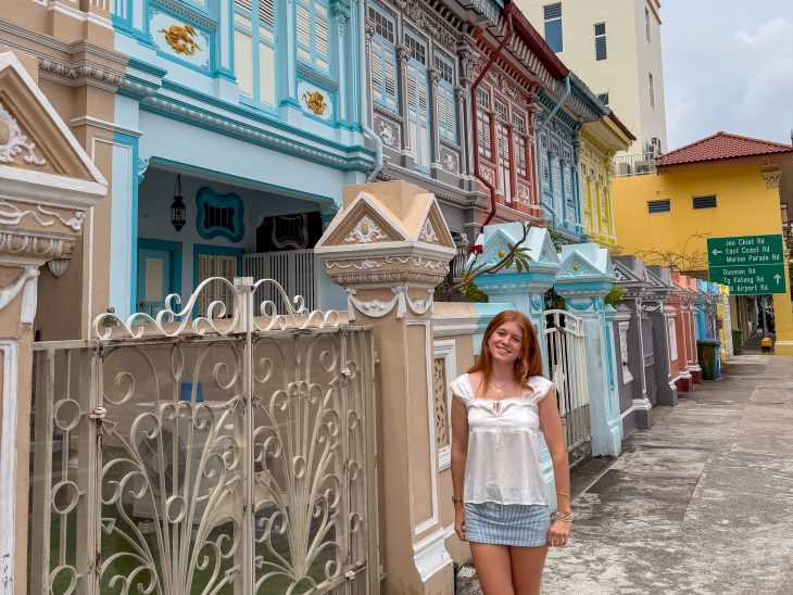 Student standing on a sidewalk beside a row of colorful historic shophouses in Singapore.