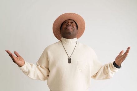 Nick Cooper wears a cream turtleneck sweater and matching wide-brim hat while looking upward with both arms raised during a portrait session against a white background.