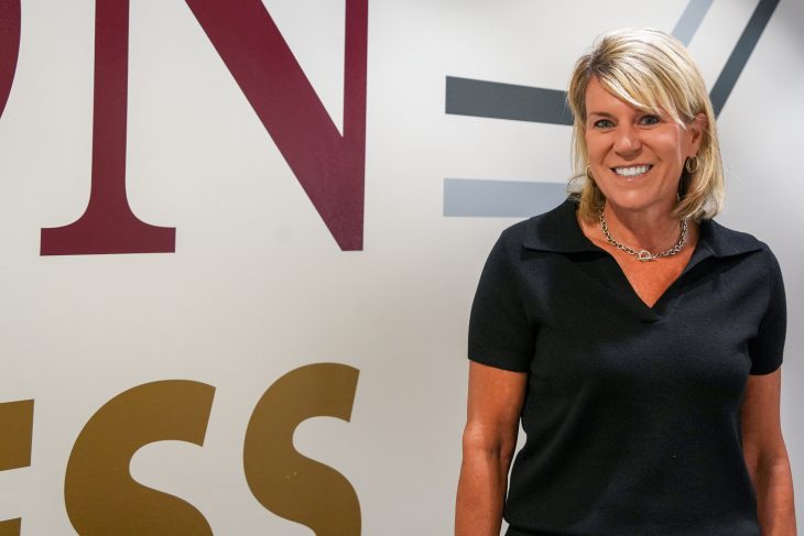 Maria Teza stands smiling in front of a wall featuring large Love School of Business branding. She is wearing a dark short-sleeve collared top, hoop earrings and a silver necklace while posing for a professional portrait.