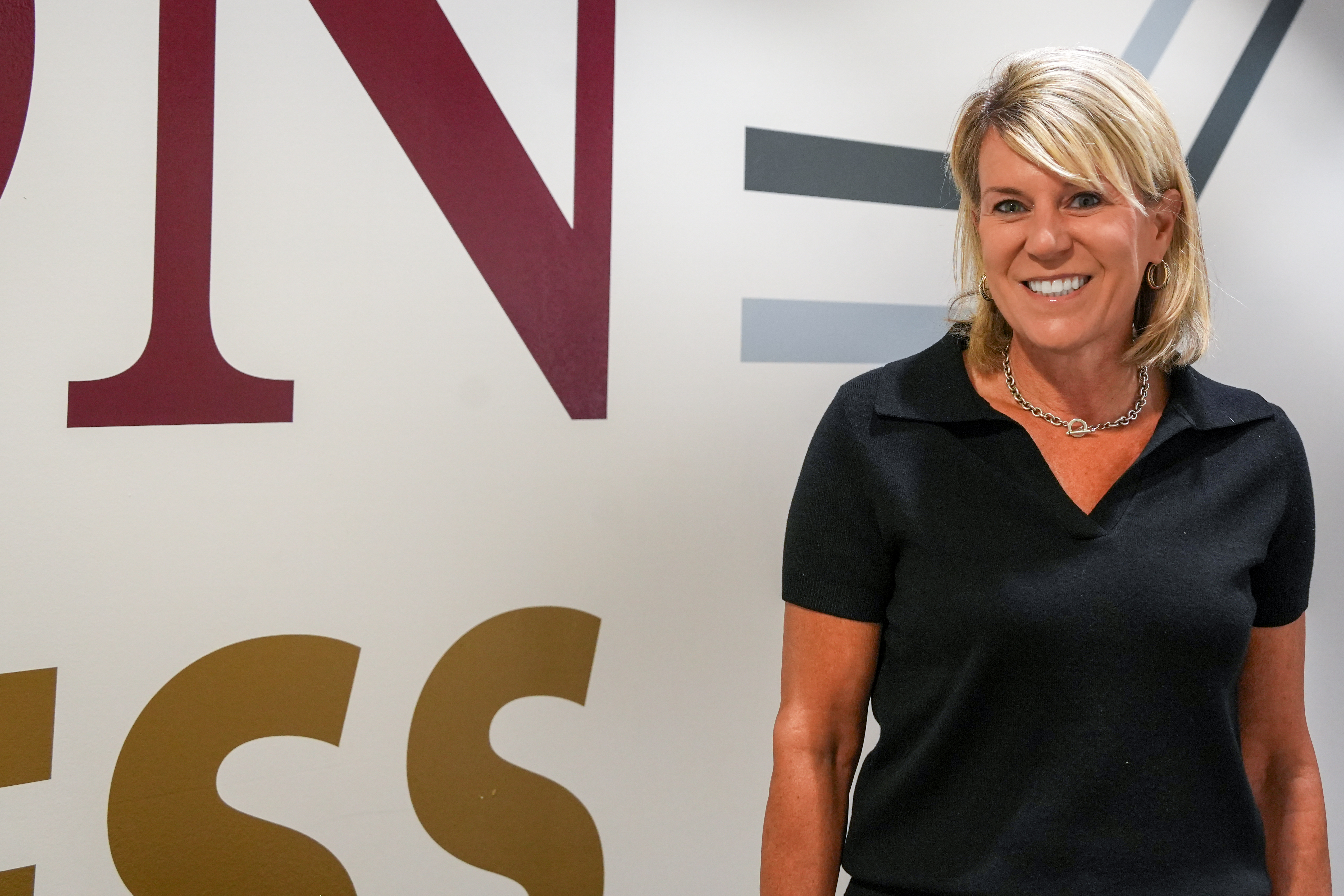 Maria Teza stands smiling in front of a wall featuring large Love School of Business branding. She is wearing a dark short-sleeve collared top, hoop earrings and a silver necklace while posing for a professional portrait.