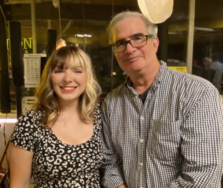 Two people pose for a photo. A woman is wearing a black and white dress. A man is wearing a blue patterned shirt. The man has glasses