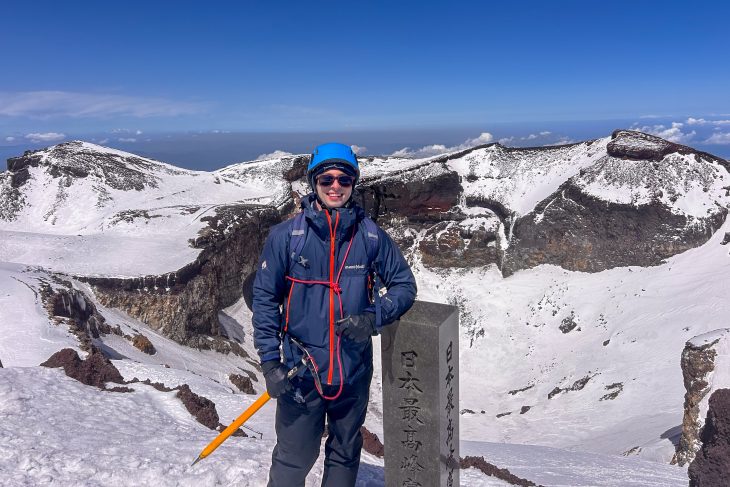 Student in winter hiking gear standing beside a summit marker on snow-covered Mount Fuji in Japan.