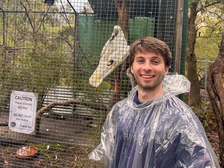 A Business Fellow in a rain poncho smiles beside a fenced enclosure with a white cockatoo perched inside during the Asia Pacific semester experience.