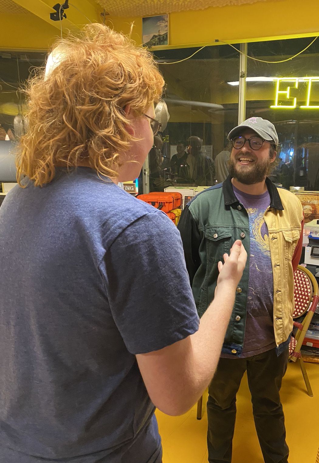 Two people chat and smile inside a brightly lit creative workspace or studio. One person gestures while speaking as the other listens, with books, equipment, and a neon Ƶ sign visible in the background.