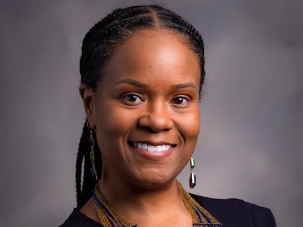 The photo shows a woman posing confidently in front of a neutral gray background. She has a warm, friendly smile and is wearing a dark, sleeveless dress. Her hair is styled in neat braids pulled back. She's adorned with a vibrant, multi-strand beaded necklace that features a mix of blue, green, red, and gold tones, and she's also wearing dangling earrings. The lighting and background suggest this is a professional portrait, possibly for business or academic use.