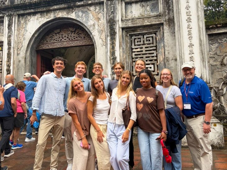 Group of Elon University Business Fellows and a faculty member standing together in front of a historic stone entrance with carved details and vertical lettering.