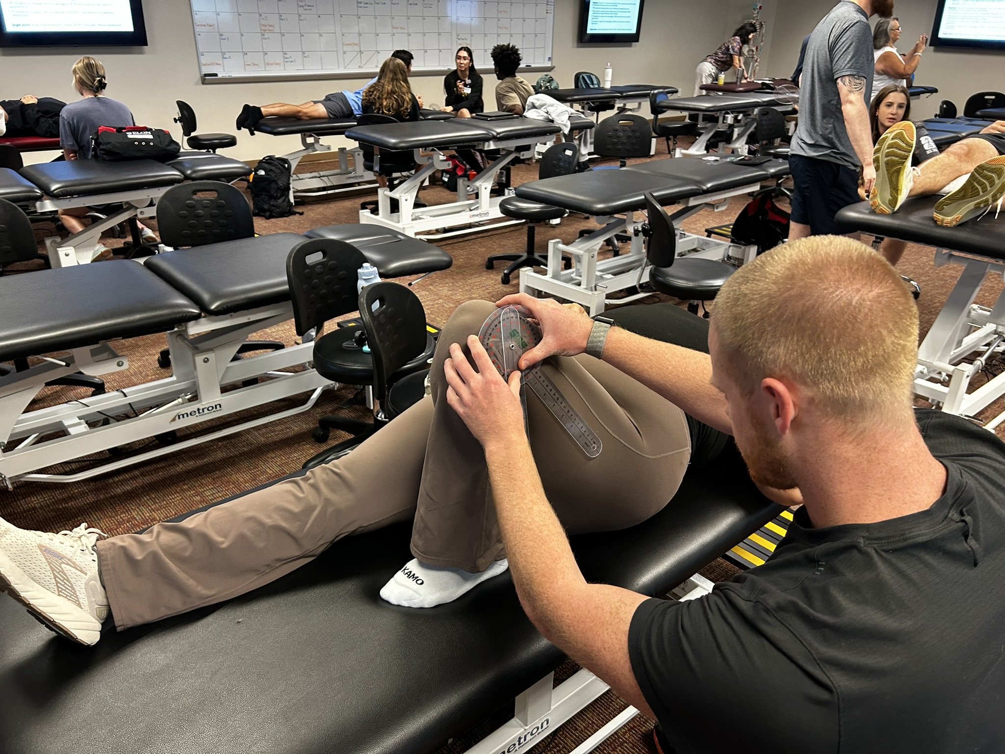 A physical therapy student measures a classmate’s knee range of motion with a goniometer during a hands-on lab while other students practice nearby.