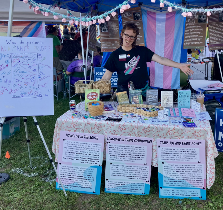 Archie Crowley at a booth during Alamance Pride. The booth is decorated with flags and has various pamphlets on display, including topics like "Trans Life in the South" and "Language in Trans Communities."