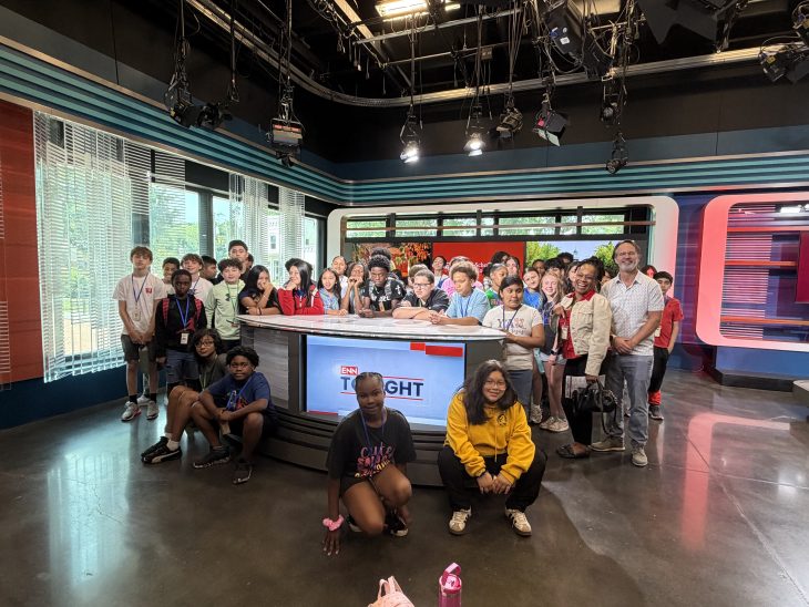 A large group of students poses behind a television news desk inside Elon University’s student media studio.