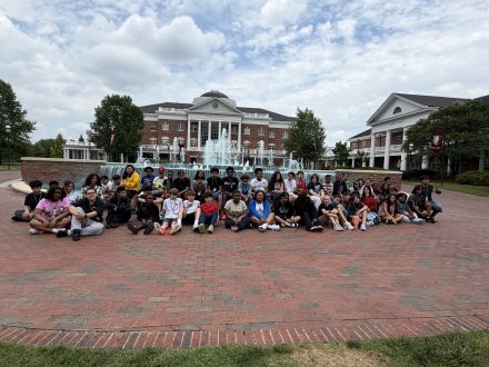 A large group of students poses in front of the fountain outside of a university brick building with a fountain during a campus visit.