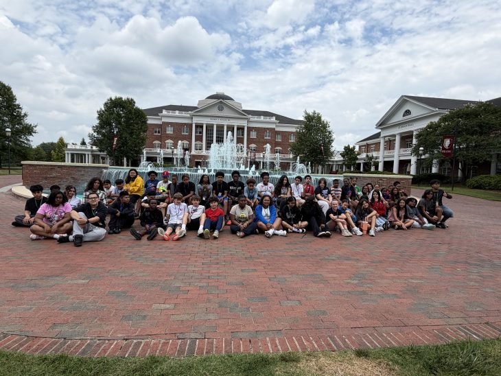 A large group of students poses in front of the fountain outside of a university brick building with a fountain during a campus visit.