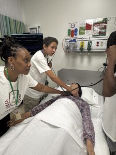 Students observe a medical simulation as an instructor demonstrates patient assessment on a mannequin in a hospital training room.