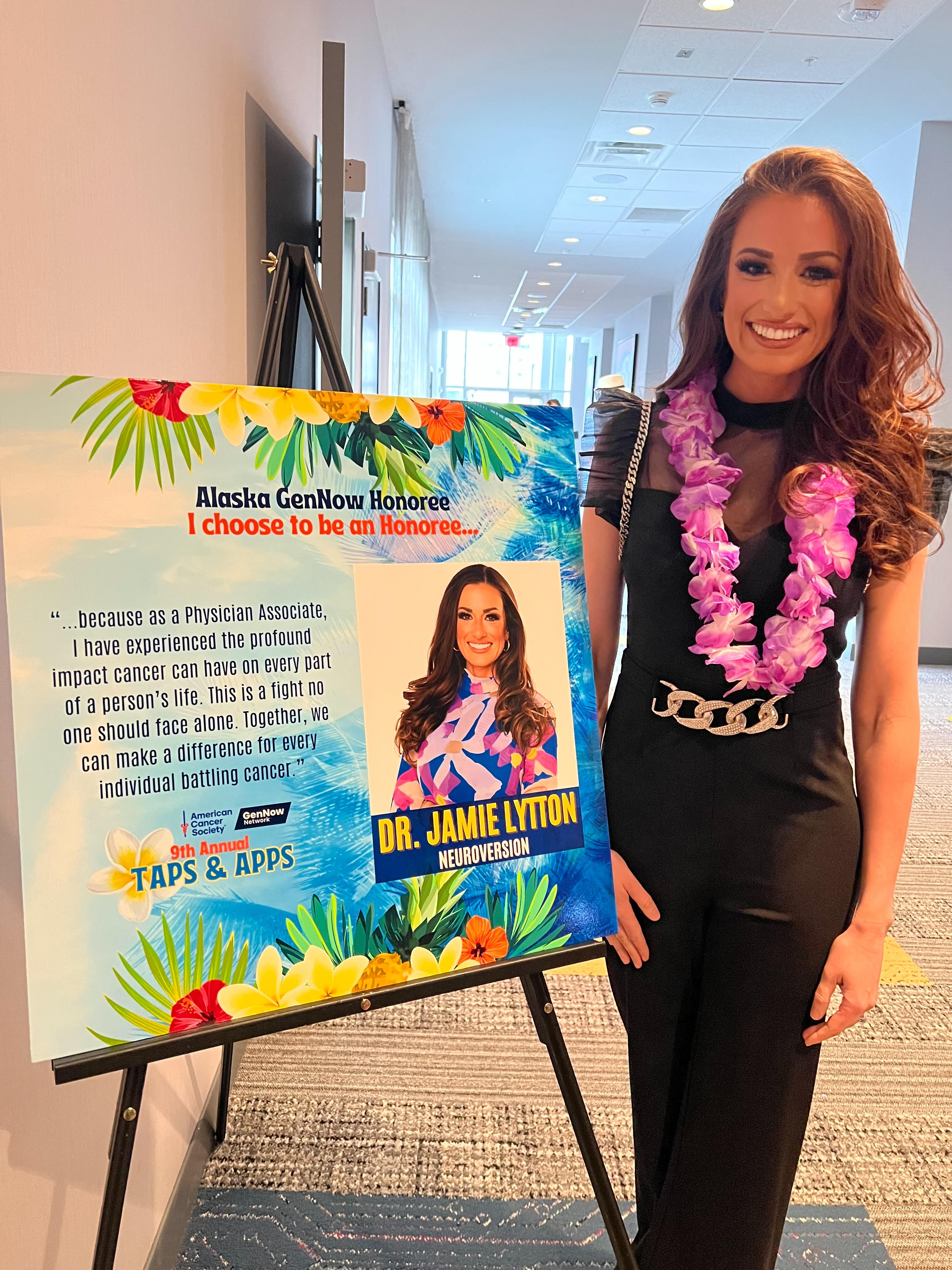 Alt text: A woman wearing a black dress and a purple flower lei smiles beside a colorful event poster recognizing her as an Alaska GenNow honoree. The poster features her photo and a message about supporting people affected by cancer through advocacy and healthcare, as part of the American Cancer Society’s 9th Annual Taps & Apps event.
