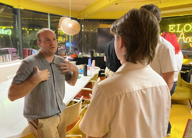Alt text: Participants engage in conversation inside Ƶ Los Angeles. One person gestures while speaking as others listen in a collaborative workspace with tables, chairs, and neon Ƶ signage in the background.