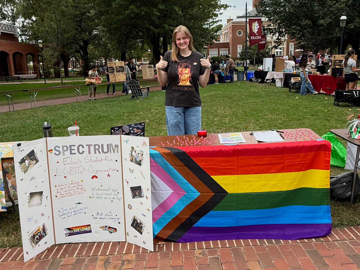 A student stands behind a table draped with a Progress Pride flag at a campus organization fair, giving two thumbs up beside an informational display.