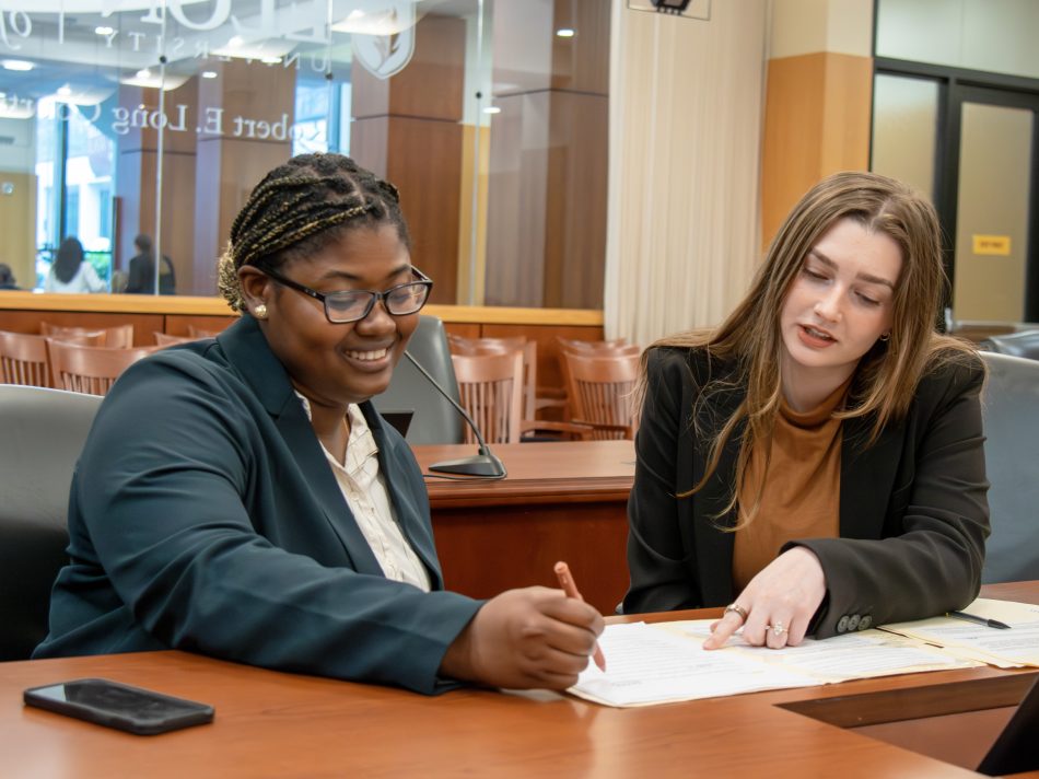 Two female law students review file folders at a desk in a courtroom. They are gesturing and discussing the notes.