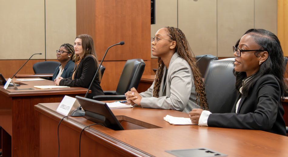 Four women shown from the right side, seated at adjacent counsel desks in a courtroom. They are smiling as they look forward.