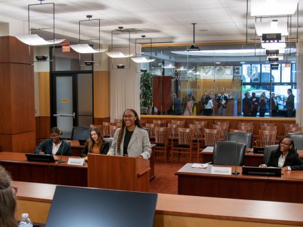 A woman addresses judges from a podium in a courtroom. Other law students are seated at adjacent tables listening.
