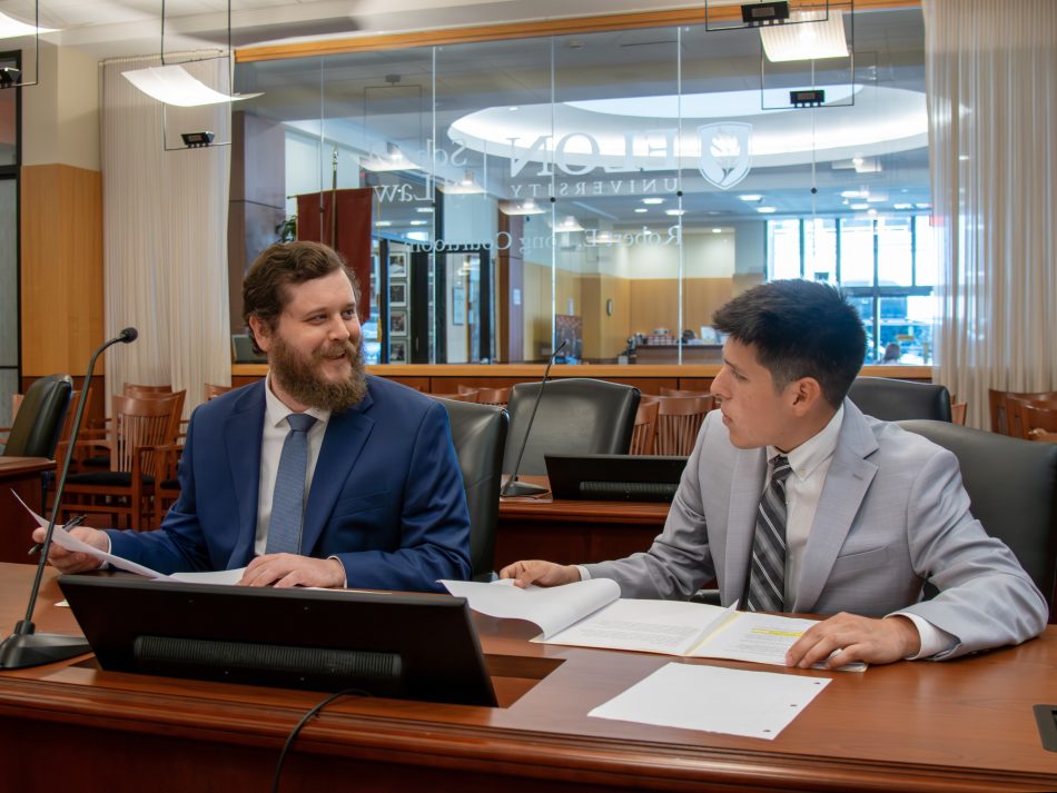 Two male students in suits and ties seated at a counsel's desk in a courtroom. They are conversing over legal documents.