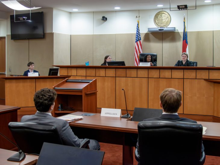 Three judges seated in a courtroom. One is speaking to students seated at counsel's table, shown from behind.