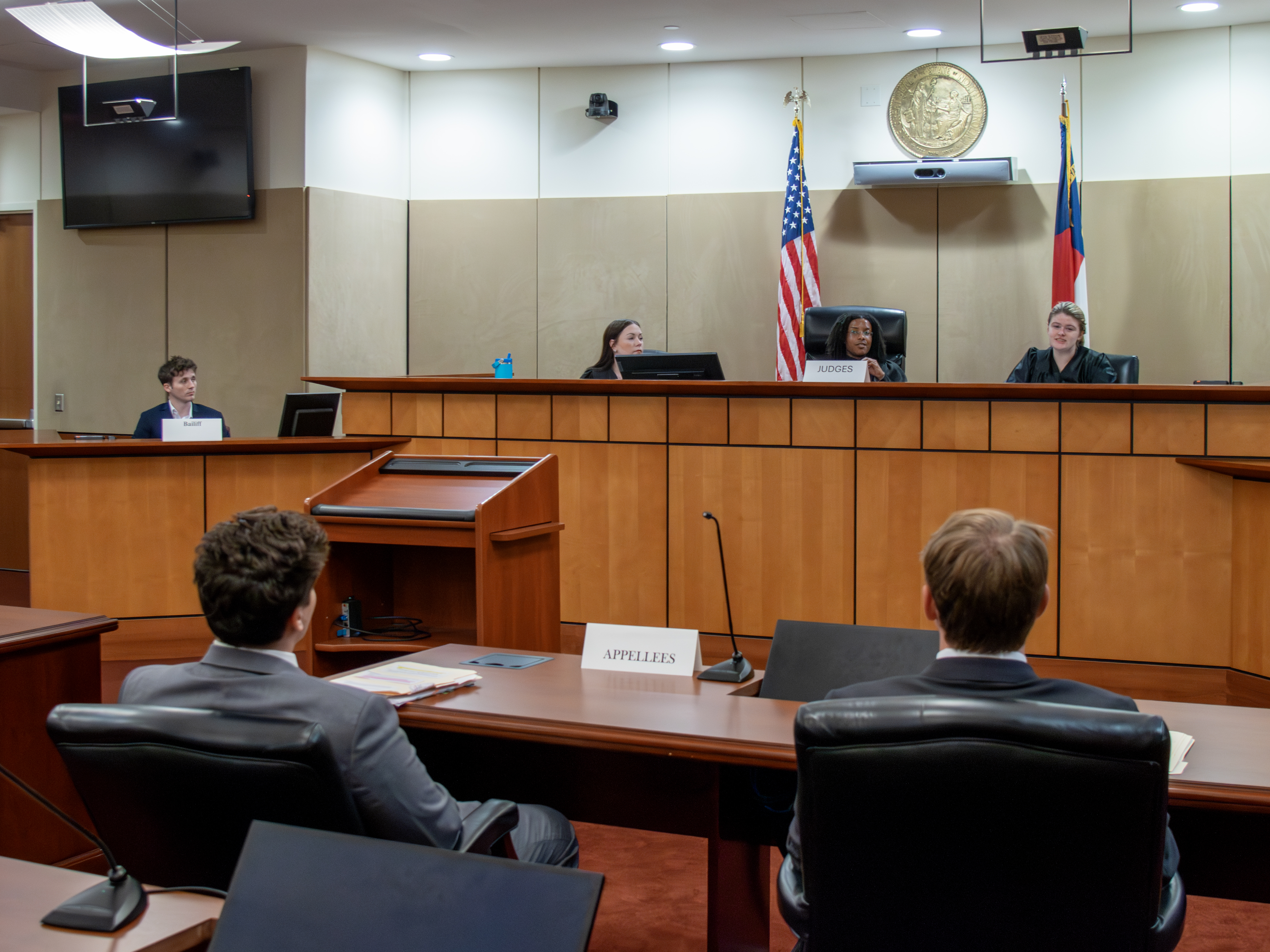 Three judges seated in a courtroom. One is speaking to students seated at counsel's table, shown from behind.