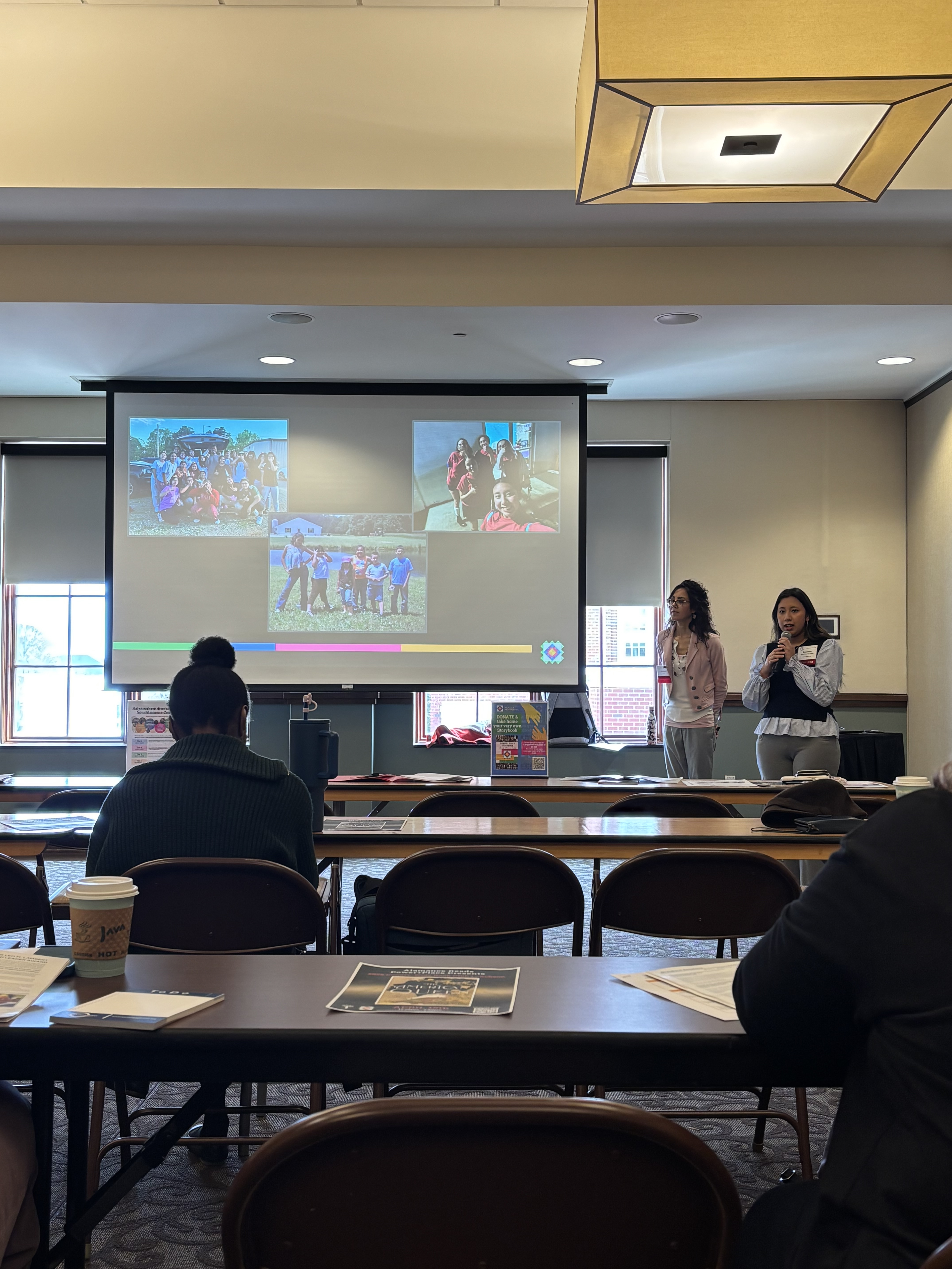 Two presenters stand at the front of a conference-style meeting room, speaking to attendees seated at tables. One presenter holds a microphone while a large projection screen behind them displays several group photos from a community or student organization activity. Coffee cups, handouts and notebooks rest on the tables as audience members listen to the presentation.