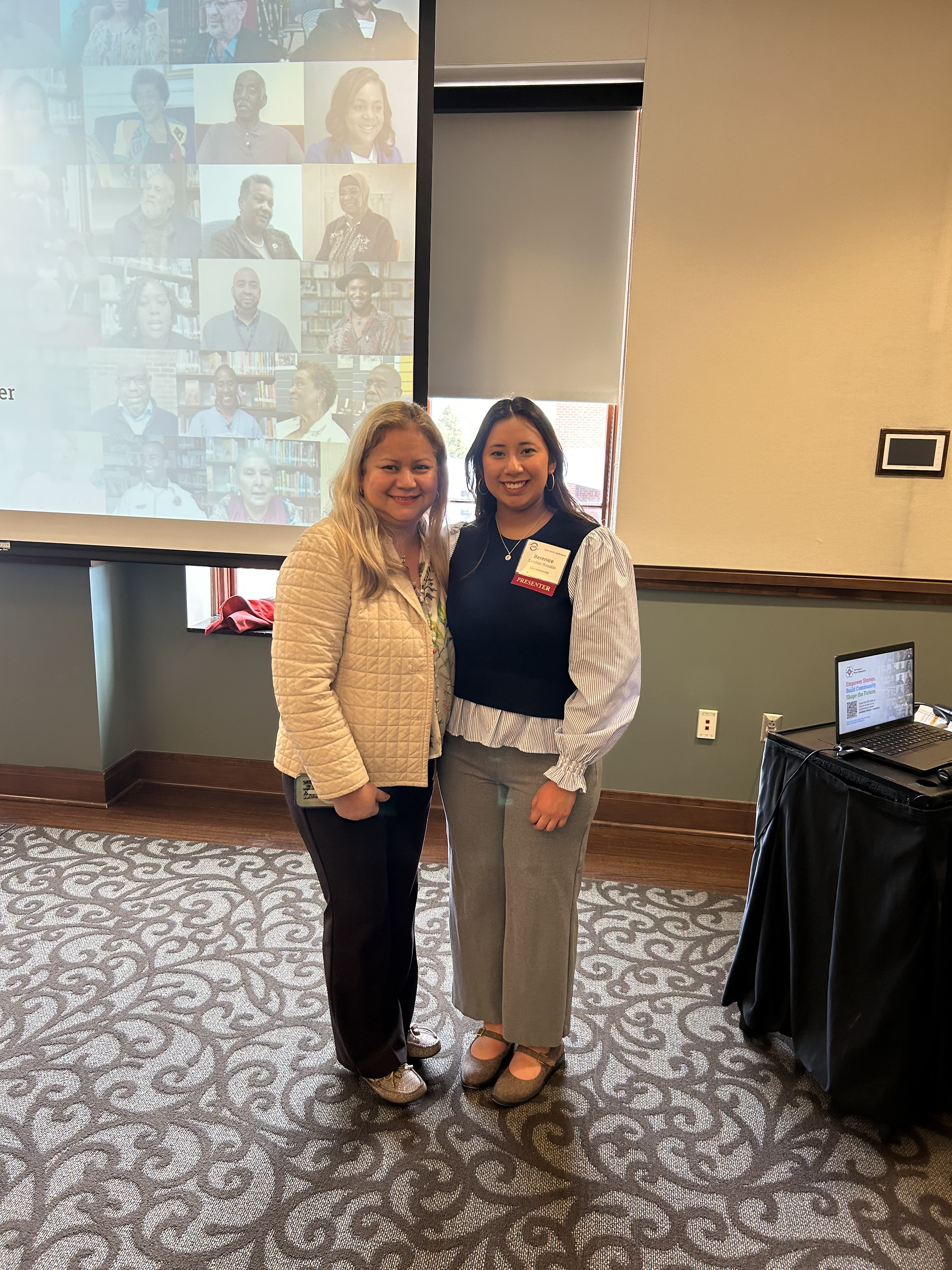 Two women stand together and smile for a photo in a conference room. Behind them, a projection screen displays a collage of portrait photos, while a laptop sits on a nearby table used for the presentation