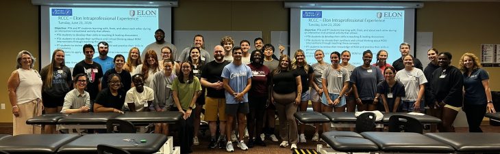 Students and faculty pose together in a physical therapy lab following an interprofessional learning session, with treatment tables and presentation screens in the background.