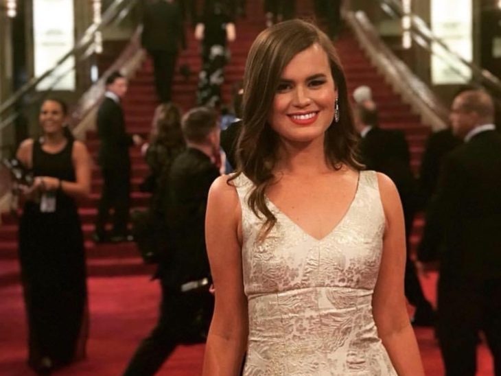 A woman in a formal gown smiles on a red carpet, with attendees and a grand staircase visible in the background.