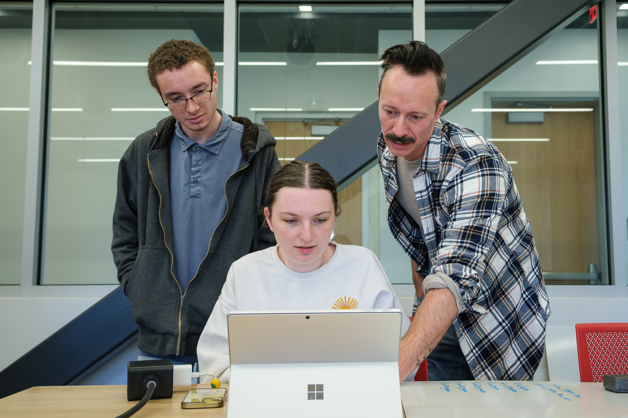 Three people stand around a laptop. Two are overlooking one person sitting down while the person on the right points to something on the screen.
