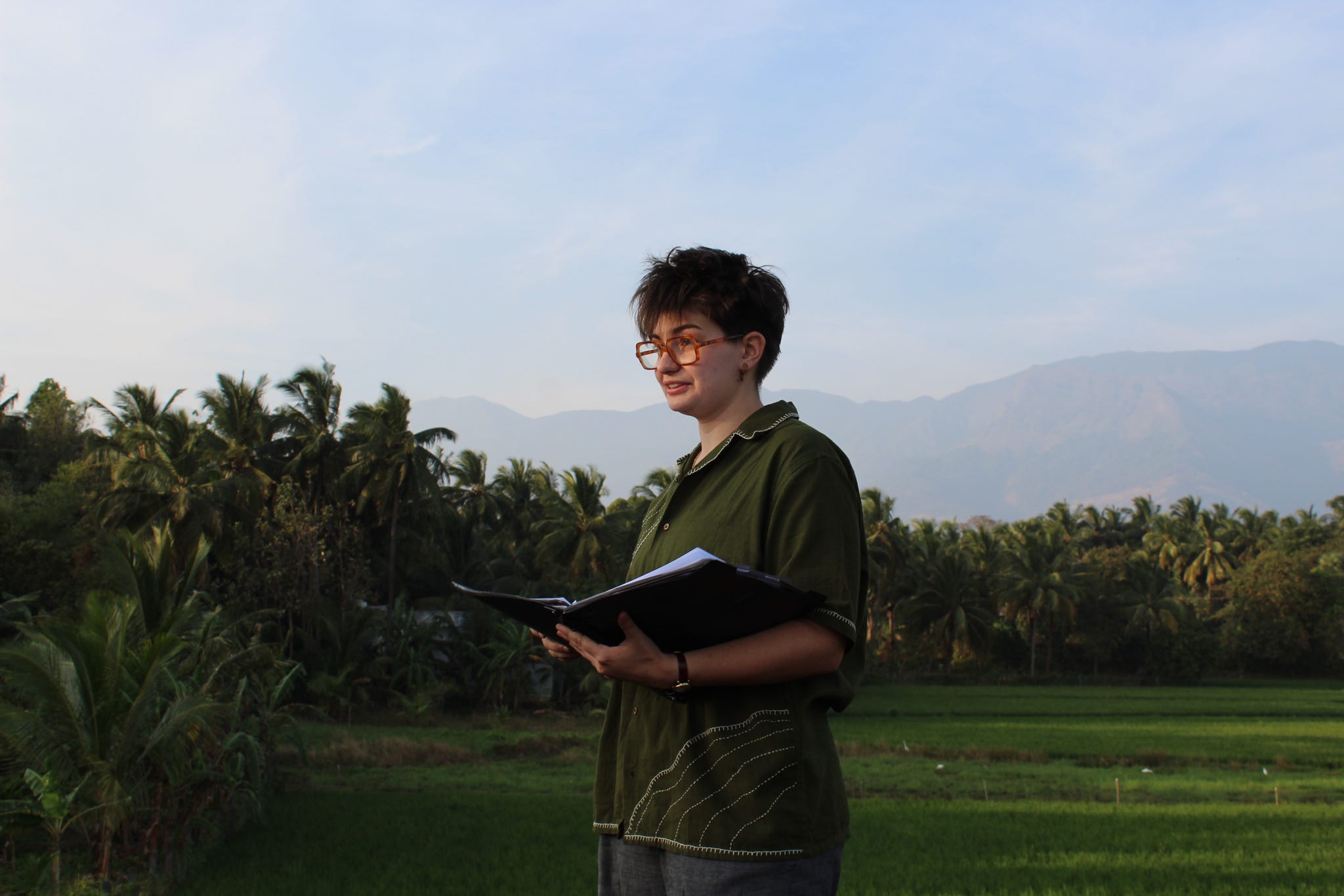 Photo of a person with a field and woods behind them, holding a book