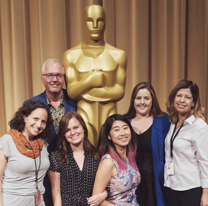 Group of six people pose beside a large Oscar statuette at a film industry event.