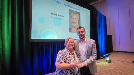 A woman and a man stand together on a conference stage, smiling while holding an award. Behind them, a large presentation screen displays the NACCU J. Paul Melanson Award, recognizing Janet Rauhe of Elon University, along with her portrait. Blue and green stage lighting frames the award presentation setting.