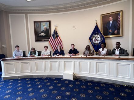 �þ��� sit behind a curved committee-style desk in a government meeting room, participating in a discussion or briefing. American and state flags stand behind them, with framed portraits displayed on the walls.