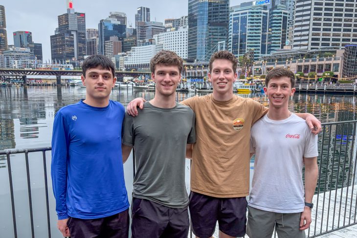 Four Business Fellows stand along the waterfront in Sydney with city buildings, boats and reflections on the harbor behind them during the Asia Pacific semester experience.