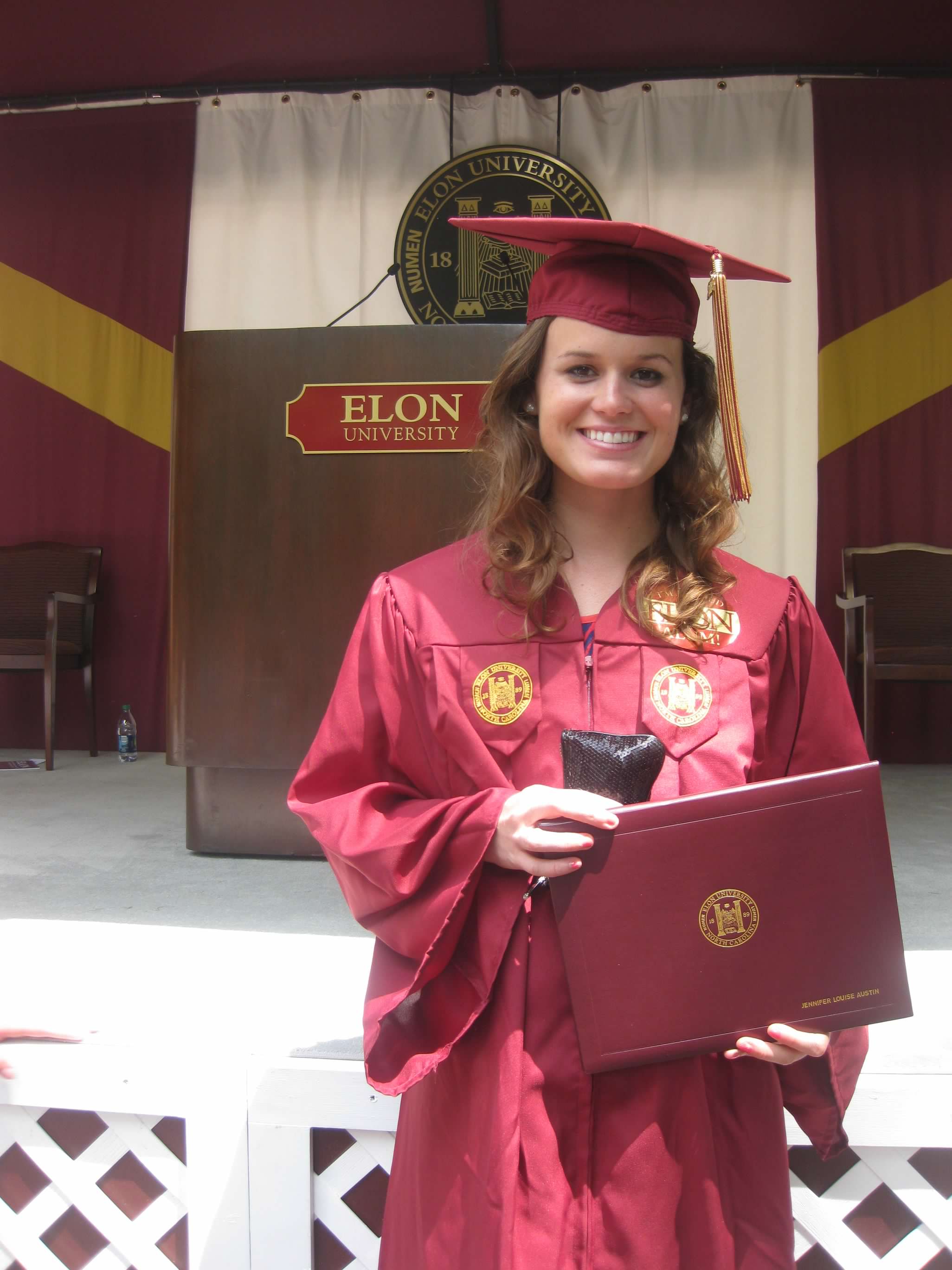 An Elon graduate in cap and gown smiles while holding a diploma folder at commencement.