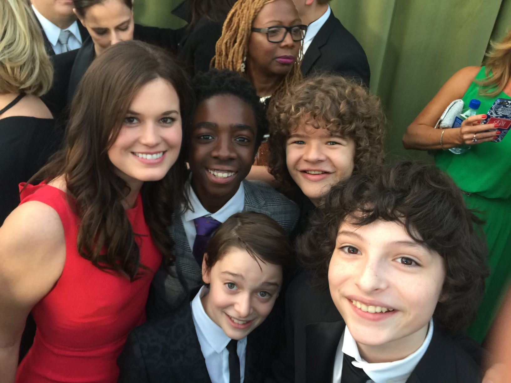 Alt text: A woman in a red dress smiles for a selfie with four young actors dressed in formal attire at an outdoor event.