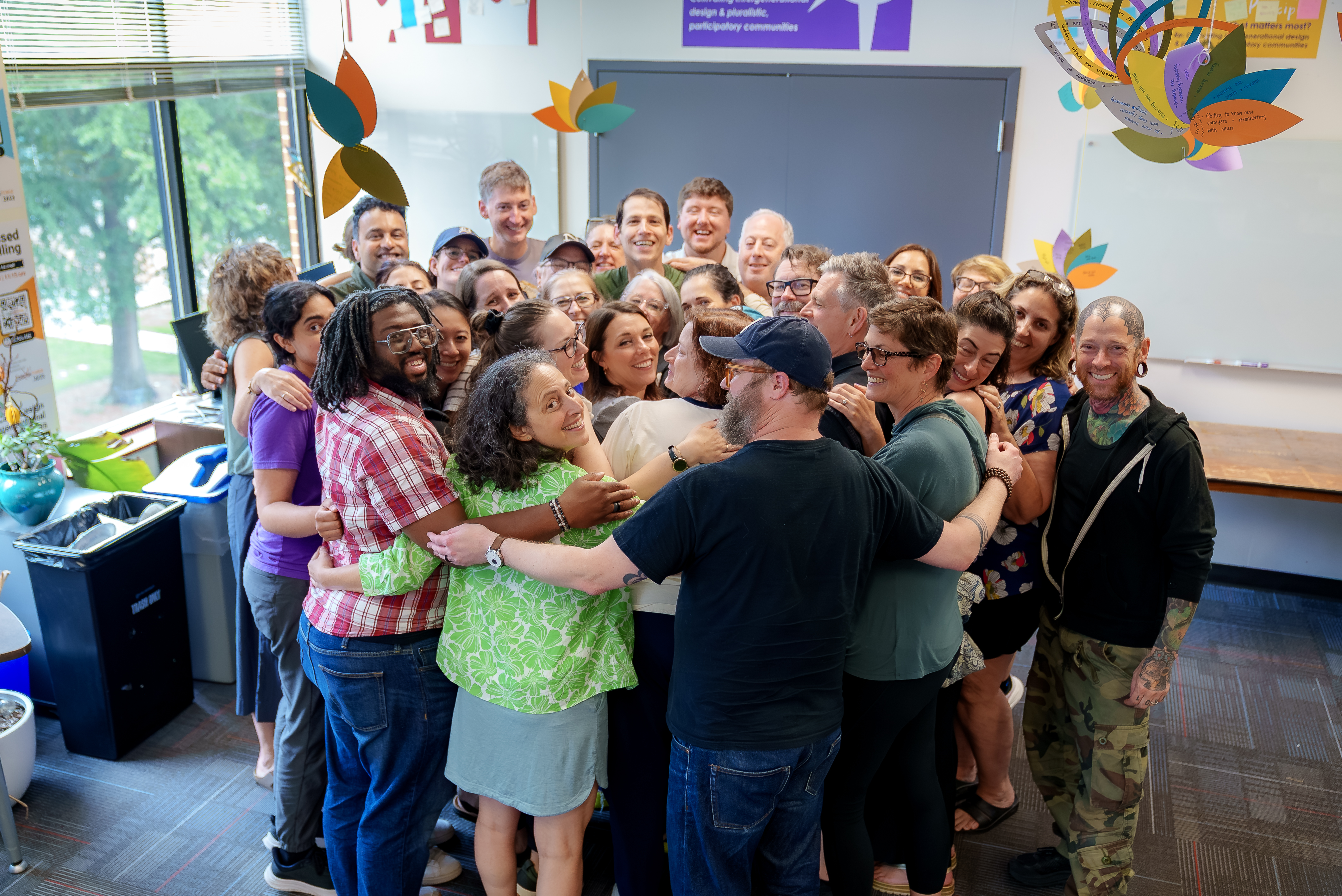 A large group of faculty and staff share a group hug inside a makerspace, celebrating the conclusion of a collaborative campus workshop or team-building event.