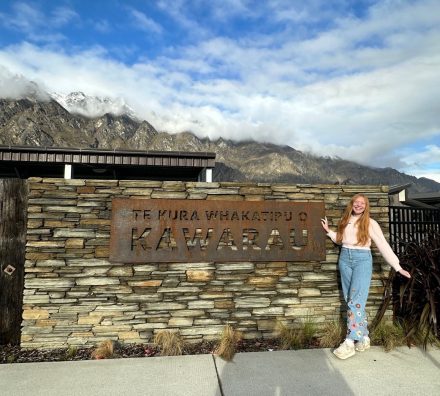 A student poses beside a sign for Te Kura Whakatipu o Kawarau in New Zealand, with snow-dusted mountains and low clouds rising in the background.
