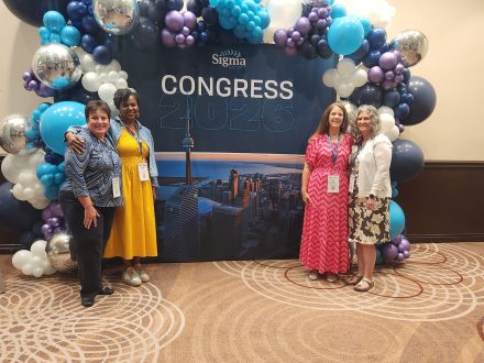 Four people pose for a photo in front of a blue sign that is bordered with blue, purple, silver and white balloons. The sign says "Sigma Conference"