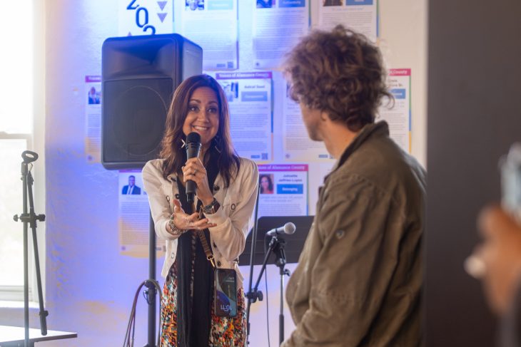 A woman speaks into a microphone while gesturing toward a seated attendee during an interactive discussion. A speaker, microphone stands and informational posters are visible in the background of the campus event space.