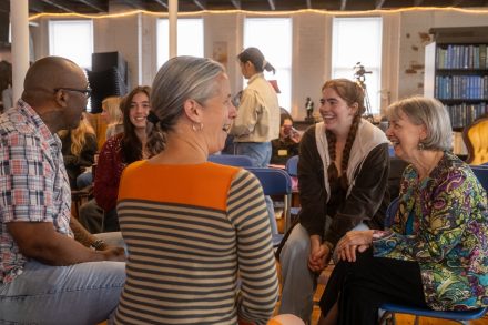 Five people sit in a circle, smiling and laughing during a group discussion in a bright campus gathering space. Bookshelves, musical instruments and other attendees in the background create a warm, informal atmosphere.