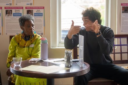 An attendee gestures while speaking to another participant seated across a small table during a campus discussion. Papers, a water bottle and informational posters are visible in the bright meeting space.