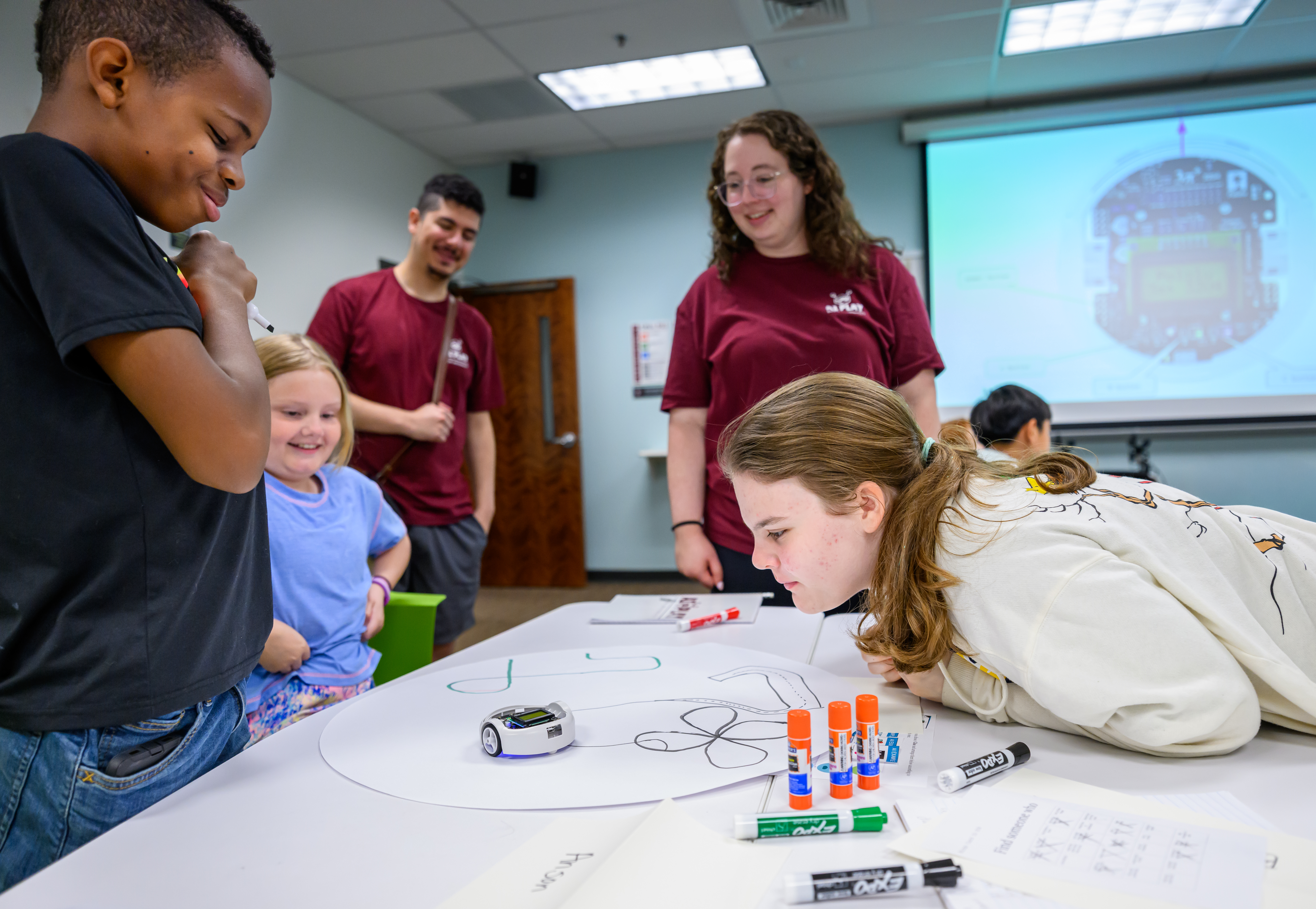 Children and student mentors gather around a table as a participant programs a small educational robot to follow a hand-drawn path during a robotics activity.