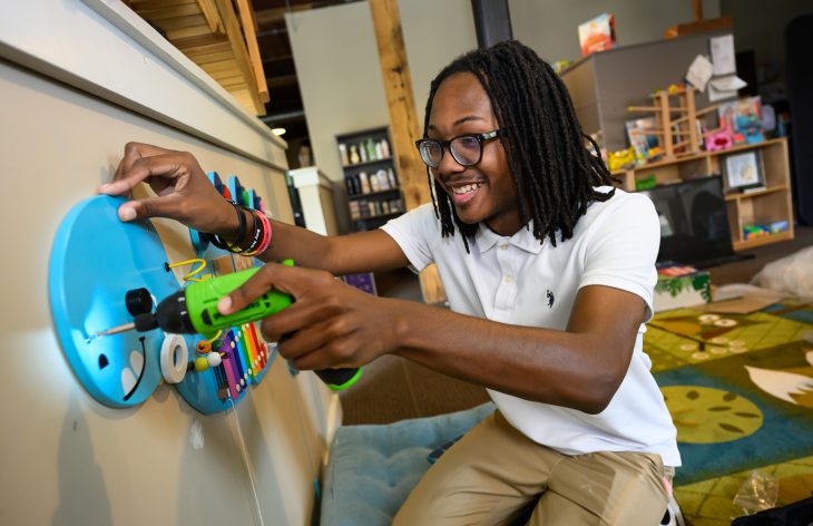 A smiling person with glasses and shoulder-length locs kneels beside a wall-mounted sensory activity board, using a green toy drill to interact with colorful gears and components in a classroom or children’s play space.