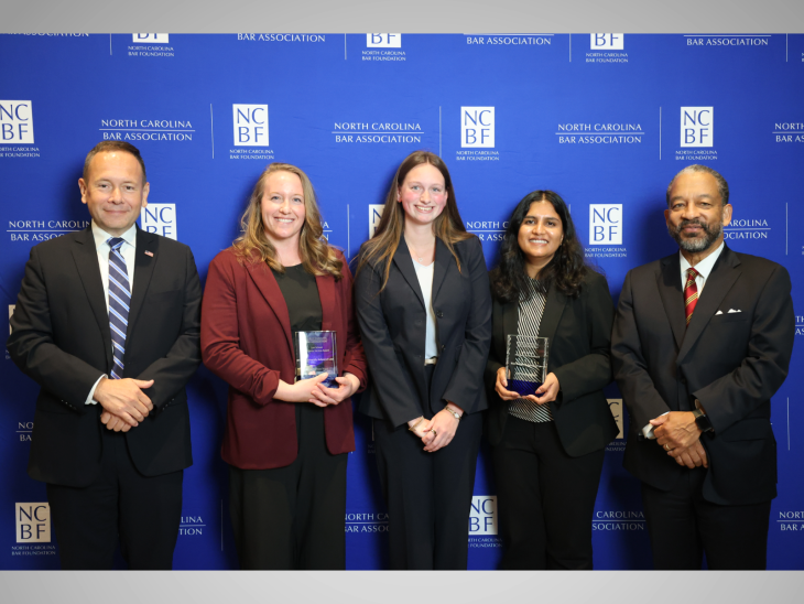 Five people standing in a row for a posed photo. Students are holiding an award.